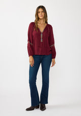 [Color: Berry] A front facing image of a brunette model wearing a burgundy boho blouse with colorful embroidered details, a split v-neckline with ties, and delicate pintuck details along the sleeve. 
