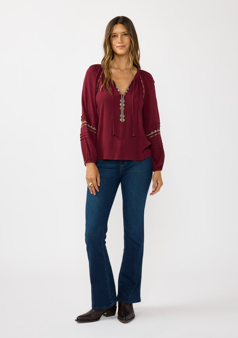 [Color: Berry] A front facing image of a brunette model wearing a burgundy boho blouse with colorful embroidered details, a split v-neckline with ties, and delicate pintuck details along the sleeve. 