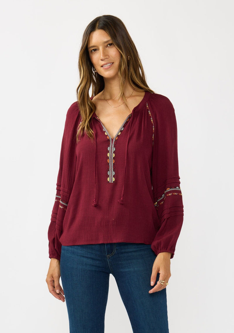 [Color: Berry] A front facing image of a brunette model wearing a burgundy boho blouse with colorful embroidered details, a split v-neckline with ties, and delicate pintuck details along the sleeve. 