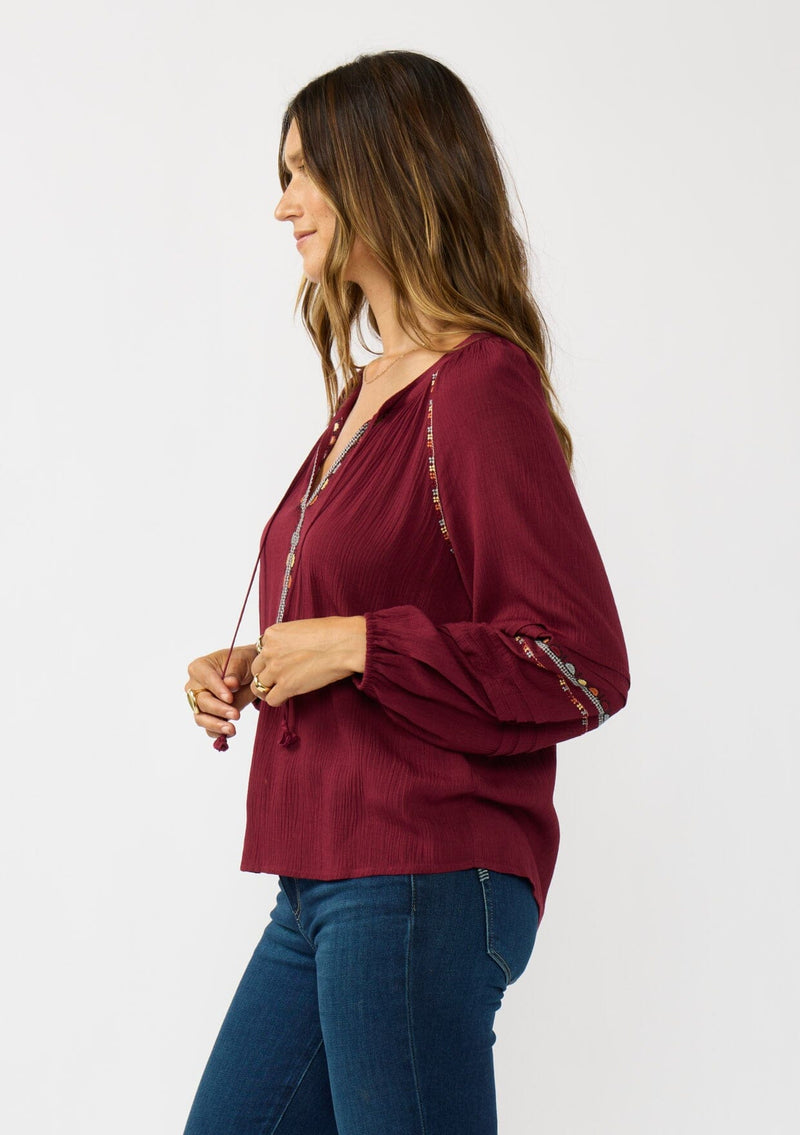 [Color: Berry] A side facing image of a brunette model wearing a burgundy boho blouse with colorful embroidered details, a split v-neckline with ties, and delicate pintuck details along the sleeve. 