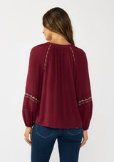 [Color: Berry] A back facing image of a brunette model wearing a burgundy boho blouse with colorful embroidered details, a split v-neckline with ties, and delicate pintuck details along the sleeve. 