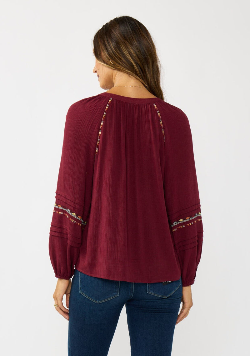 [Color: Berry] A back facing image of a brunette model wearing a burgundy boho blouse with colorful embroidered details, a split v-neckline with ties, and delicate pintuck details along the sleeve. 