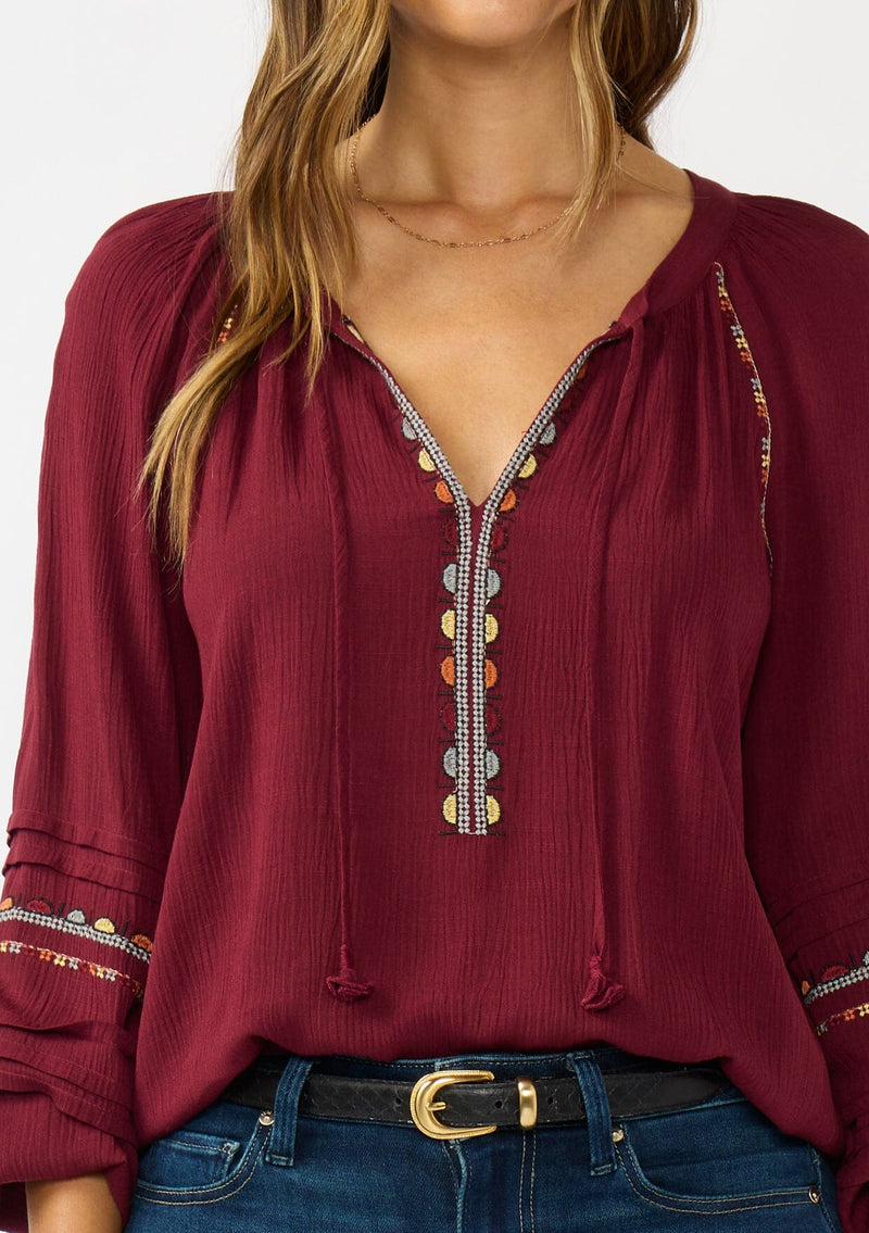 [Color: Berry] A detail image of a brunette model wearing a burgundy boho blouse with colorful embroidered details, a split v-neckline with ties, and delicate pintuck details along the sleeve. 