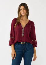 [Color: Berry] A front facing image of a brunette model wearing a burgundy boho blouse with colorful embroidered details, a split v-neckline with ties, and delicate pintuck details along the sleeve. 