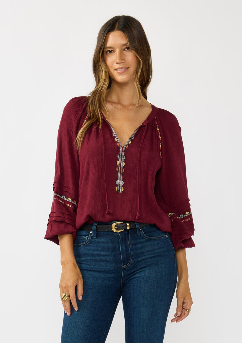 [Color: Berry] A front facing image of a brunette model wearing a burgundy boho blouse with colorful embroidered details, a split v-neckline with ties, and delicate pintuck details along the sleeve. 