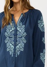 [Color: Navy] A detail image of a brunette model wearing a blue embroidered blouse with a button front and long sleeves with button cuffs. A boho blouse for the fall season. 