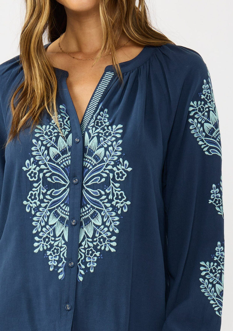 [Color: Navy] A detail image of a brunette model wearing a blue embroidered blouse with a button front and long sleeves with button cuffs. A boho blouse for the fall season. 