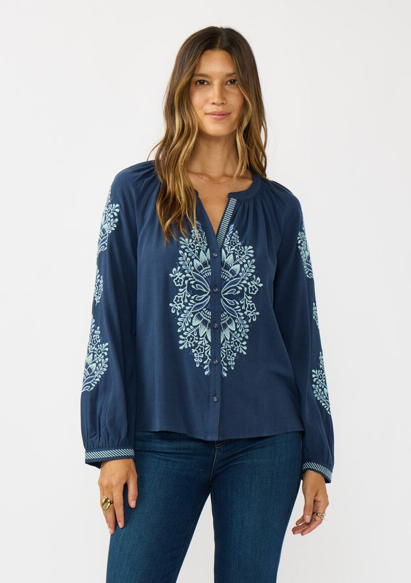 [Color: Navy] A front facing image of a brunette model wearing a blue embroidered blouse with a button front and long sleeves with button cuffs. A boho blouse for the fall season. 