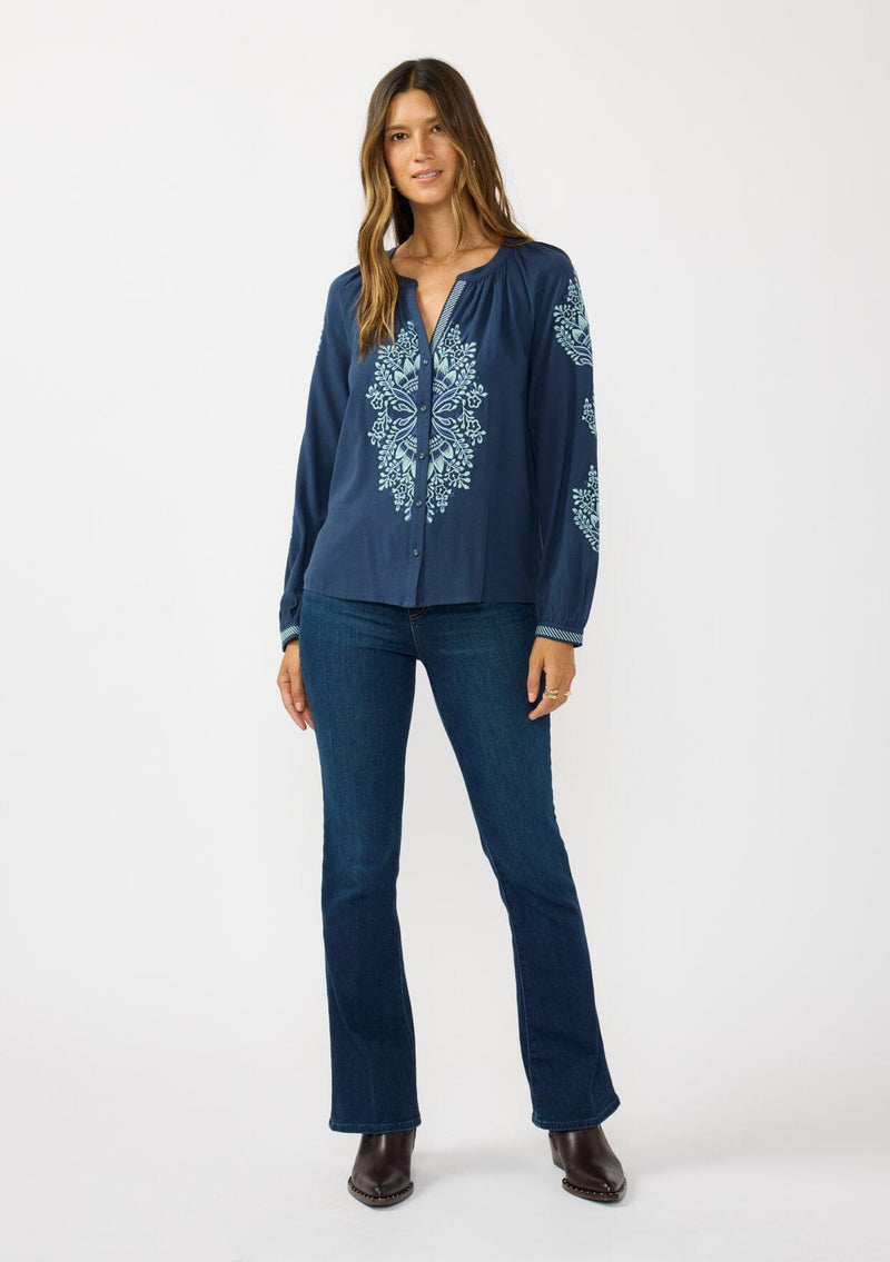 [Color: Navy] A front facing image of a brunette model wearing a blue embroidered blouse with a button front and long sleeves with button cuffs. A boho blouse for the fall season. 