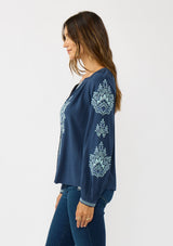 [Color: Navy] A side facing image of a brunette model wearing a blue embroidered blouse with a button front and long sleeves with button cuffs. A boho blouse for the fall season. 