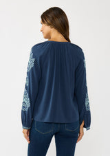 [Color: Navy] A back facing image of a brunette model wearing a blue embroidered blouse with a button front and long sleeves with button cuffs. A boho blouse for the fall season. 