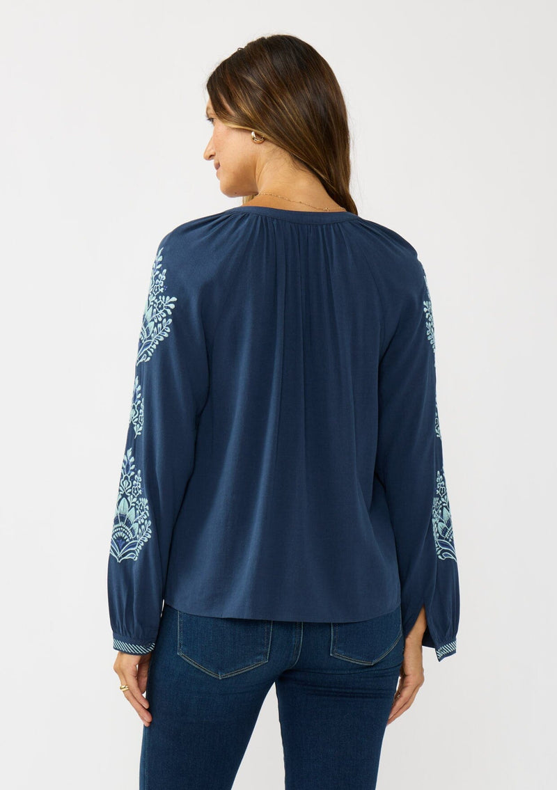 [Color: Navy] A back facing image of a brunette model wearing a blue embroidered blouse with a button front and long sleeves with button cuffs. A boho blouse for the fall season. 