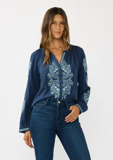 [Color: Navy] A front facing image of a brunette model wearing a blue embroidered blouse with a button front and long sleeves with button cuffs. A boho blouse for the fall season. 