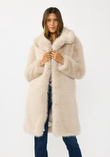 [Color: Sand] A front facing image of a brunette model wearing a beige faux fur coat with a shawl collared neckline, and side pockets. A large fur coat for the winter, holidays, and other special occasions. 