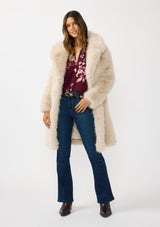 [Color: Sand] A front facing image of a brunette model wearing a beige faux fur coat with a shawl collared neckline, and side pockets. A large fur coat for the winter, holidays, and other special occasions. 