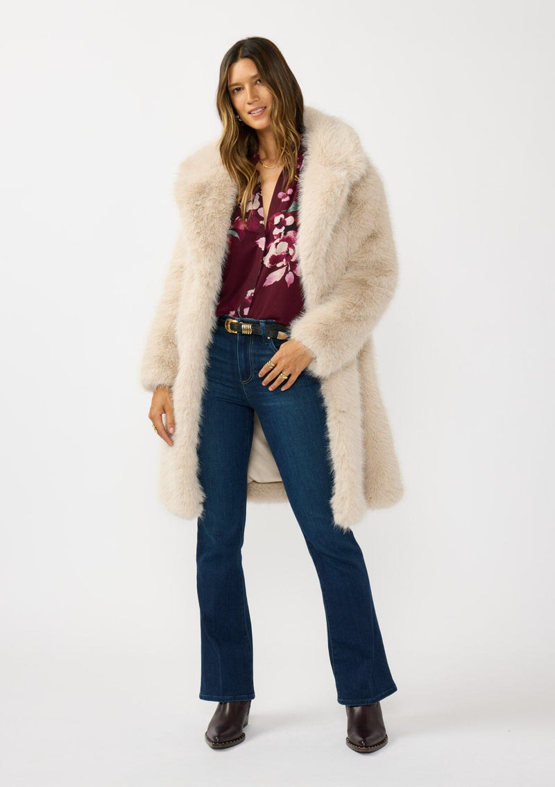 [Color: Sand] A front facing image of a brunette model wearing a beige faux fur coat with a shawl collared neckline, and side pockets. A large fur coat for the winter, holidays, and other special occasions. 