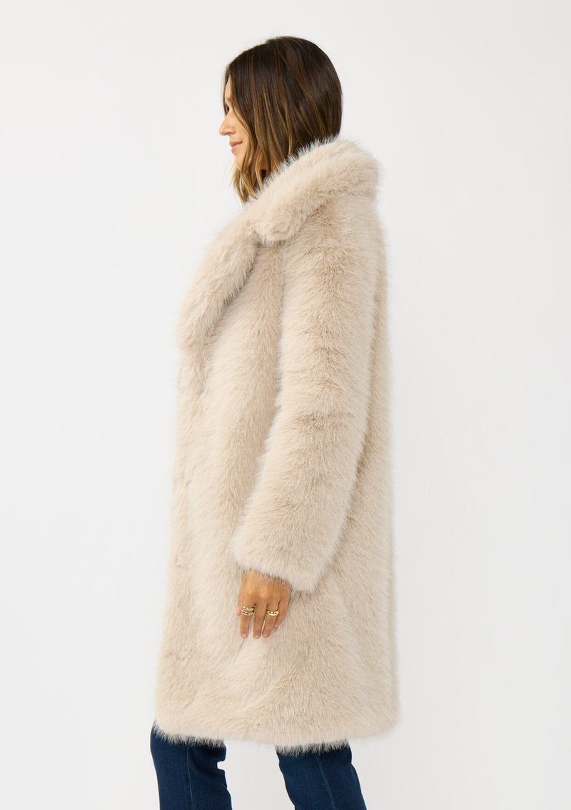 [Color: Sand] A side facing image of a brunette model wearing a beige faux fur coat with a shawl collared neckline, and side pockets. A large fur coat for the winter, holidays, and other special occasions. 