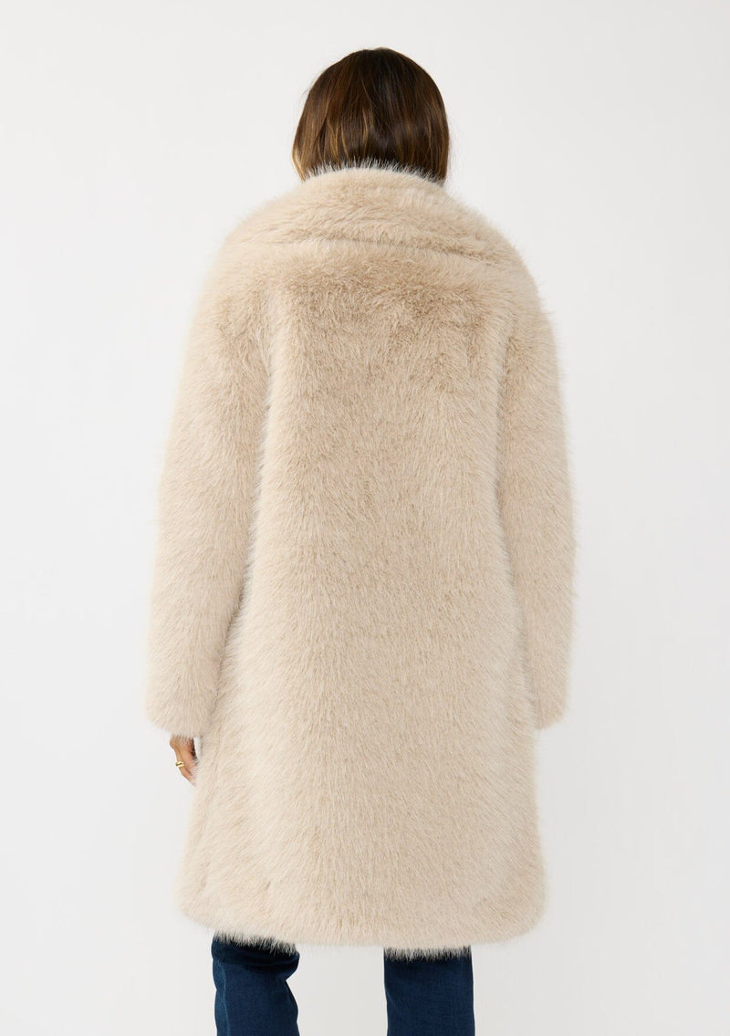 [Color: Sand] A back facing image of a brunette model wearing a beige faux fur coat with a shawl collared neckline, and side pockets. A large fur coat for the winter, holidays, and other special occasions. 