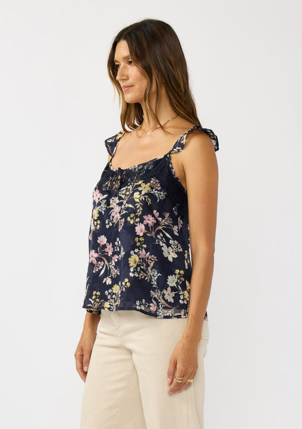 [Color: Navy/Yellow] A side facing image a navy floral top with burnout details. Featuring an adjustable straps with ruffle detail, a scoop neckline with tie detail, and lace trim. A feminine and romantic top for the fall season. 