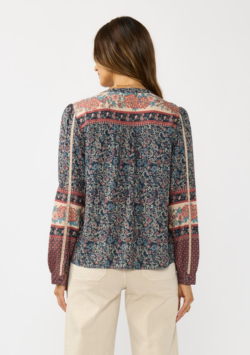 [Color: Navy/Rose] A back facing image of a brunette model wearing a navy floral border print blouse with a button front and long sleeves with lace trim accents.