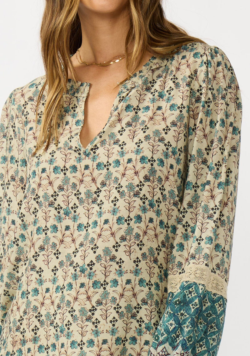 [Color: Taupe/Dusty Teal] A detail image of a brunette model wearing a green teal floral blouse with a split v-neckline, lace accent on sleeves, and ruffled elastic cuffs. A fall boho blouse for casual styling. 