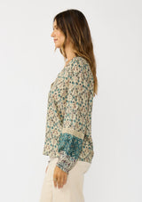 [Color: Taupe/Dusty Teal] A side facing image of a brunette model wearing a green teal floral blouse with a split v-neckline, lace accent on sleeves, and ruffled elastic cuffs. A fall boho blouse for casual styling. 