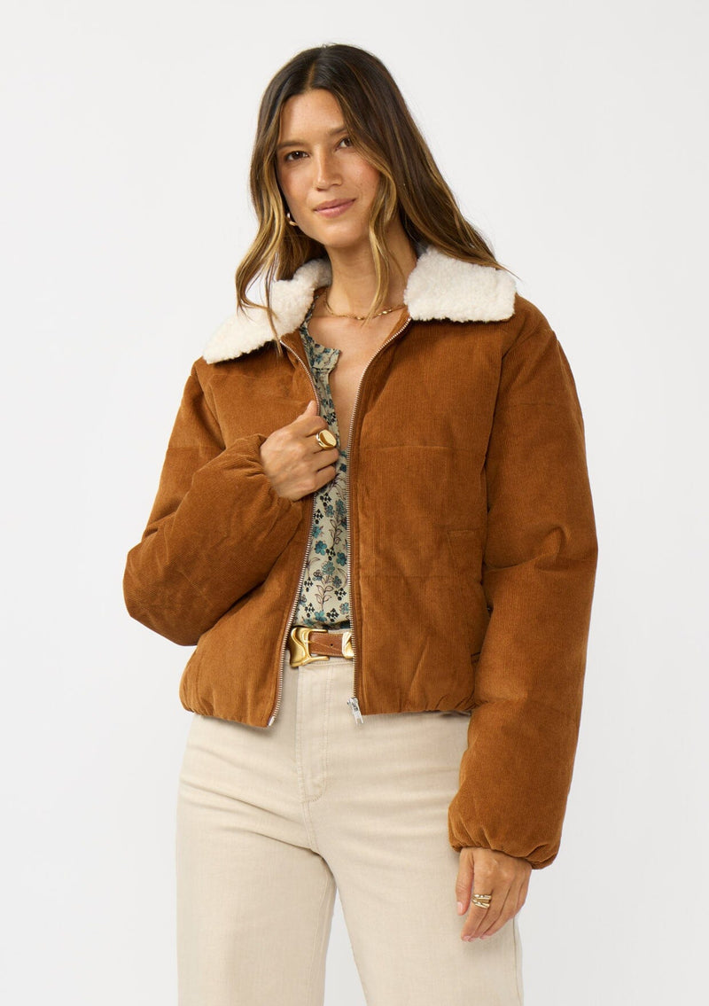 [Color: Camel] A front facing image of a brunette model wearing a brown corduroy puffer jacket with a sherpa collar, zip-up front, and welt pockets. 