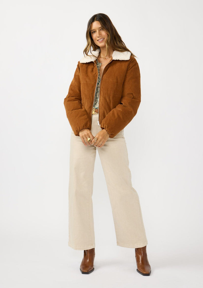 [Color: Camel] A front facing image of a brunette model wearing a brown corduroy puffer jacket with a sherpa collar, zip-up front, and welt pockets. 