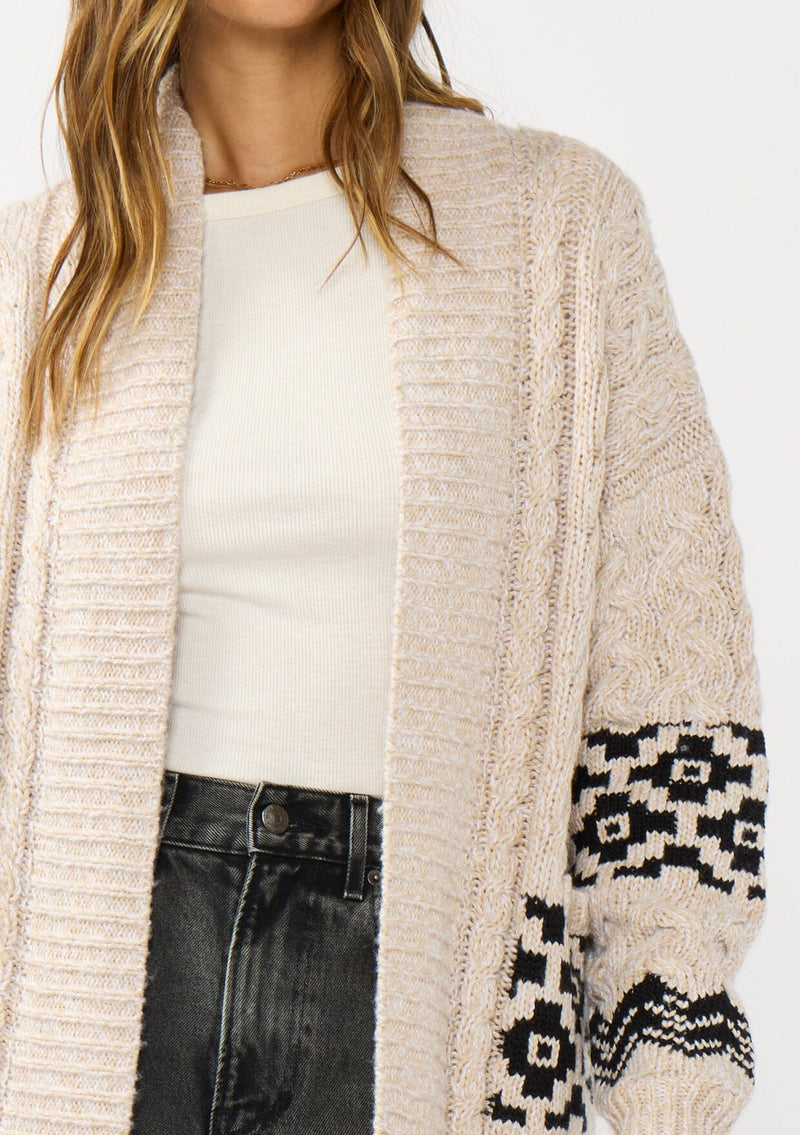 [Color: Natural/Black] A detail image of a brunette model wearing a marled knit cardigan with a trendy diamond pattern, an open front, and ribbed border. A southwestern inspired knit cardigan with a boho relaxed fit. 