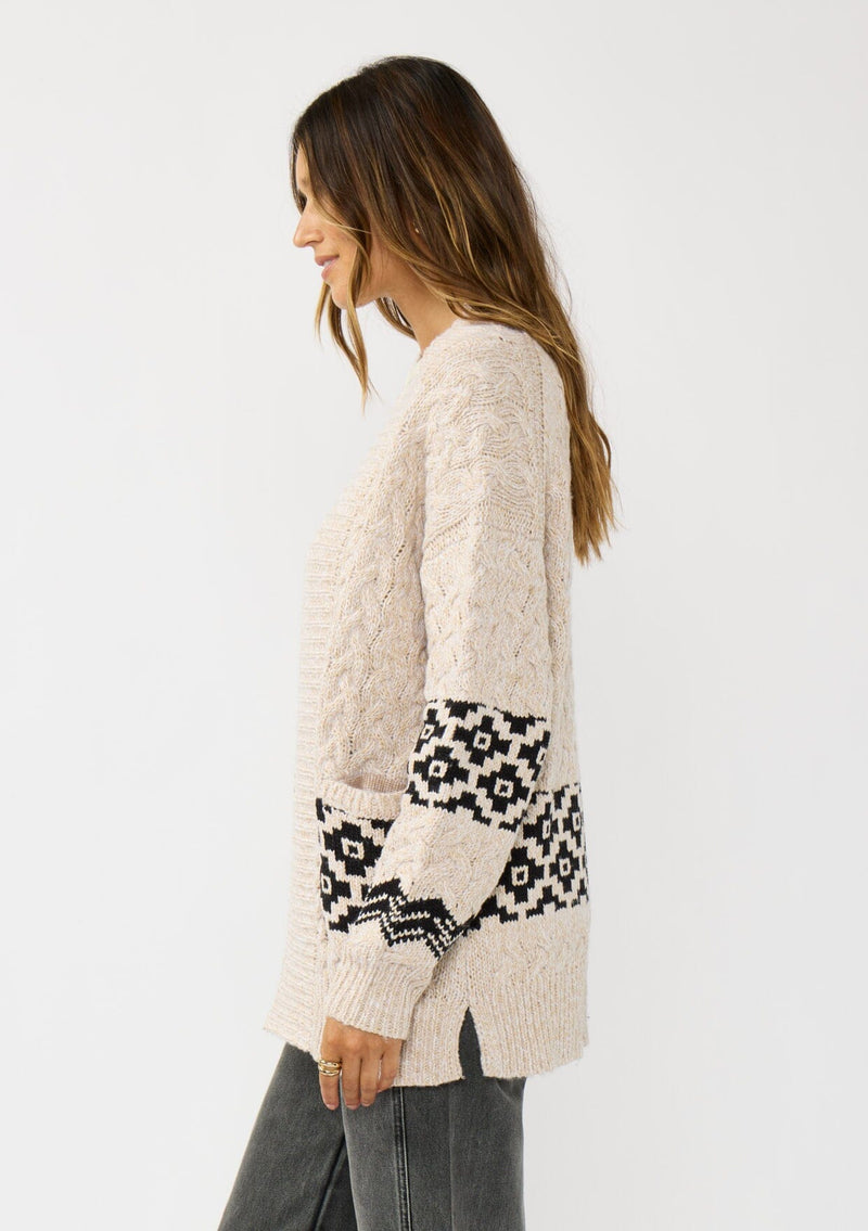 [Color: Natural/Black] A side facing image of a brunette model wearing a marled knit cardigan with a trendy diamond pattern, an open front, and ribbed border. A southwestern inspired knit cardigan with a boho relaxed fit. 