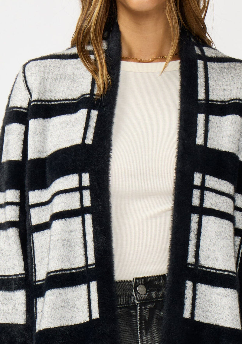 [Color: Black/Grey] A detail image of a brunette model wearing a black and grey fuzzy plaid cardigan with an open front and patch pockets. A cozy cardigan for the fall season. 