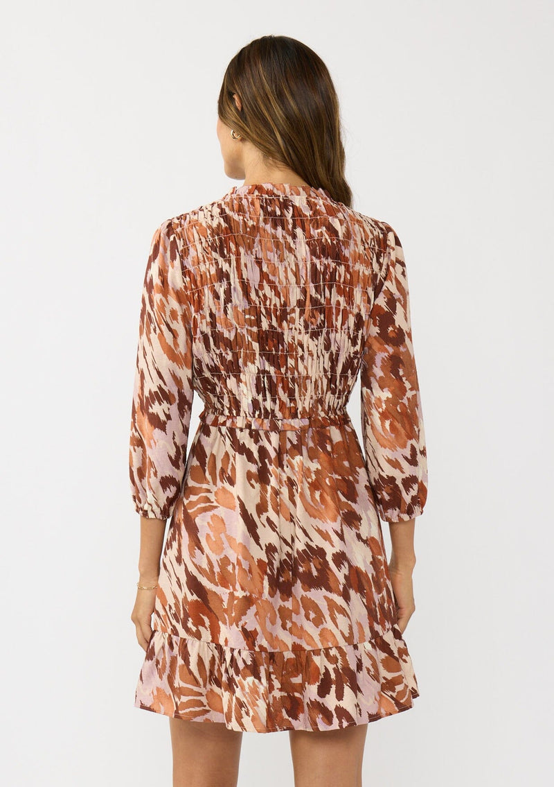 [Color: Nude/Brown] A back facing image of a brunette model wearing a brown abstract print mini dress with a smocked top, long sleeves, and a ruffle trimmed hem. A fall boho dress for casual outings. 