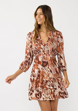 [Color: Nude/Brown] A front facing image of a brunette model wearing a brown abstract print mini dress with a smocked top, long sleeves, and a ruffle trimmed hem. A fall boho dress for casual outings. 