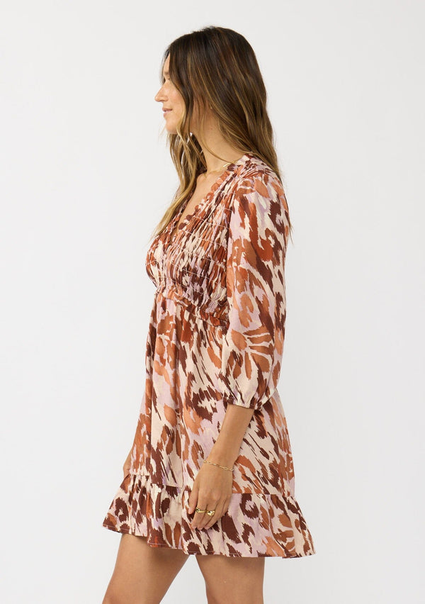 [Color: Nude/Brown] A side facing image of a brunette model wearing a brown abstract print mini dress with a smocked top, long sleeves, and a ruffle trimmed hem. A fall boho dress for casual outings. 