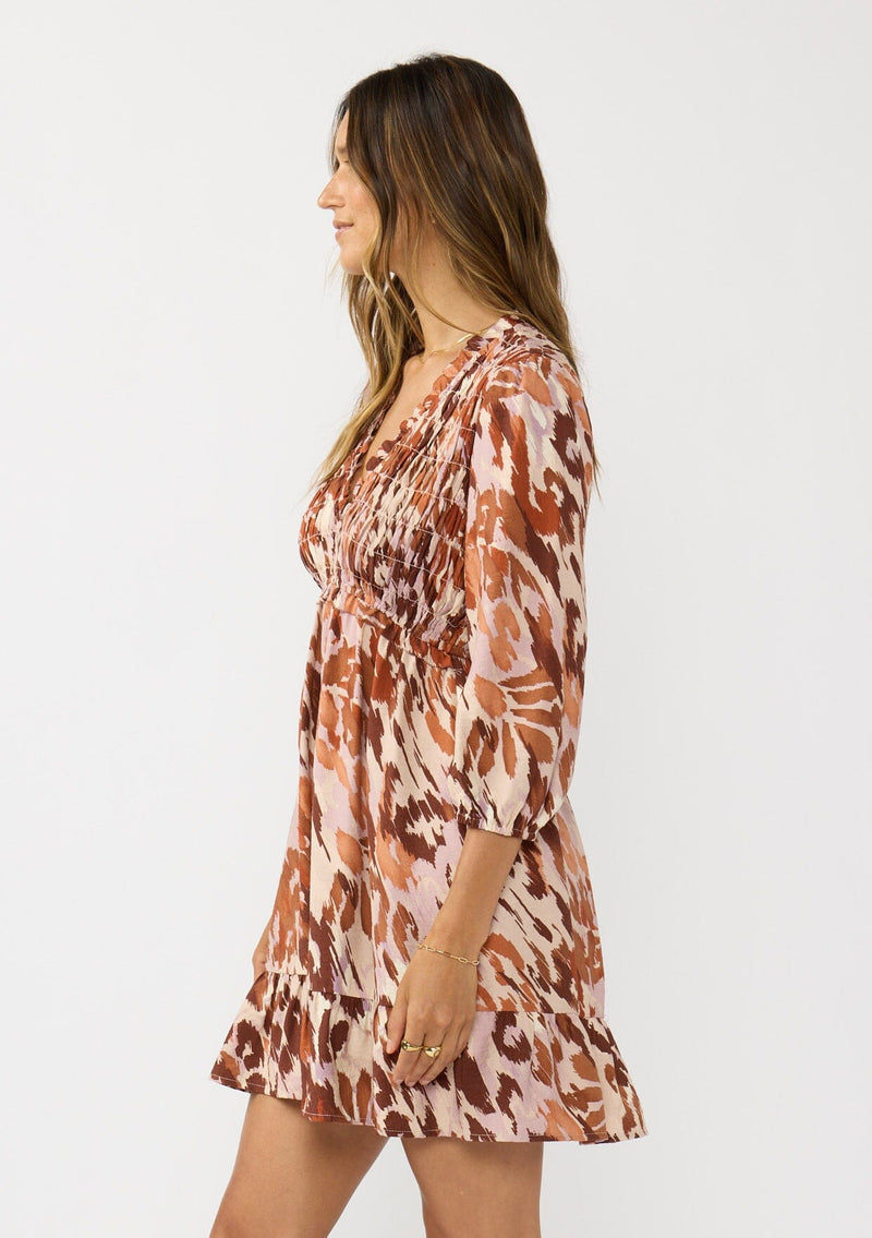 [Color: Nude/Brown] A side facing image of a brunette model wearing a brown abstract print mini dress with a smocked top, long sleeves, and a ruffle trimmed hem. A fall boho dress for casual outings. 