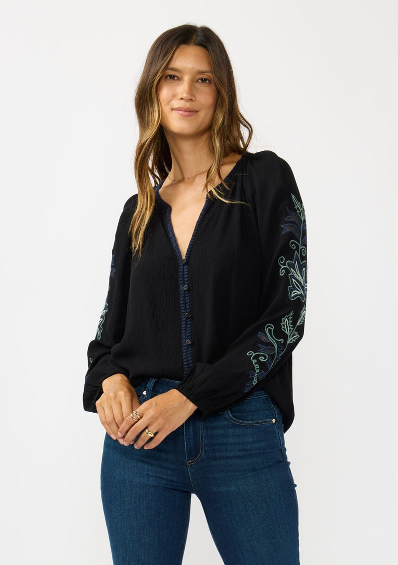 [Color: Black] A front facing image of a brunette model wearing a flowy black blouse with colorful embroidered sleeve details, a button front, and blanket stitch detailing. Perfect for fall styling. 