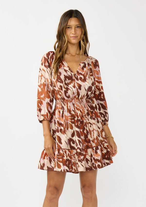 [Color: Nude/Brown] A front facing image of a brunette model wearing a brown abstract print mini dress with a split v-neckline, smocked waist, and 3/4 length sleeves. A fall boho mini dress for casual outings. 