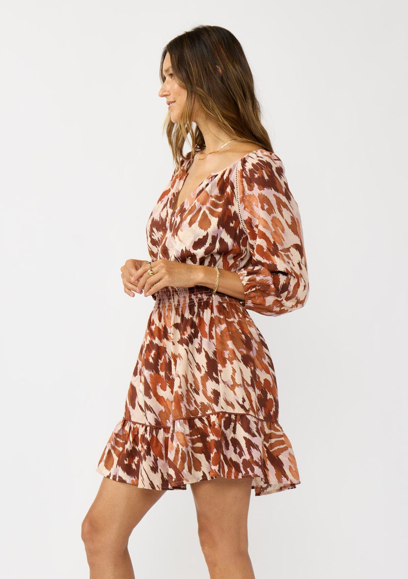 [Color: Nude/Brown] A side facing image of a brunette model wearing a brown abstract print mini dress with a split v-neckline, smocked waist, and 3/4 length sleeves. A fall boho mini dress for casual outings. 