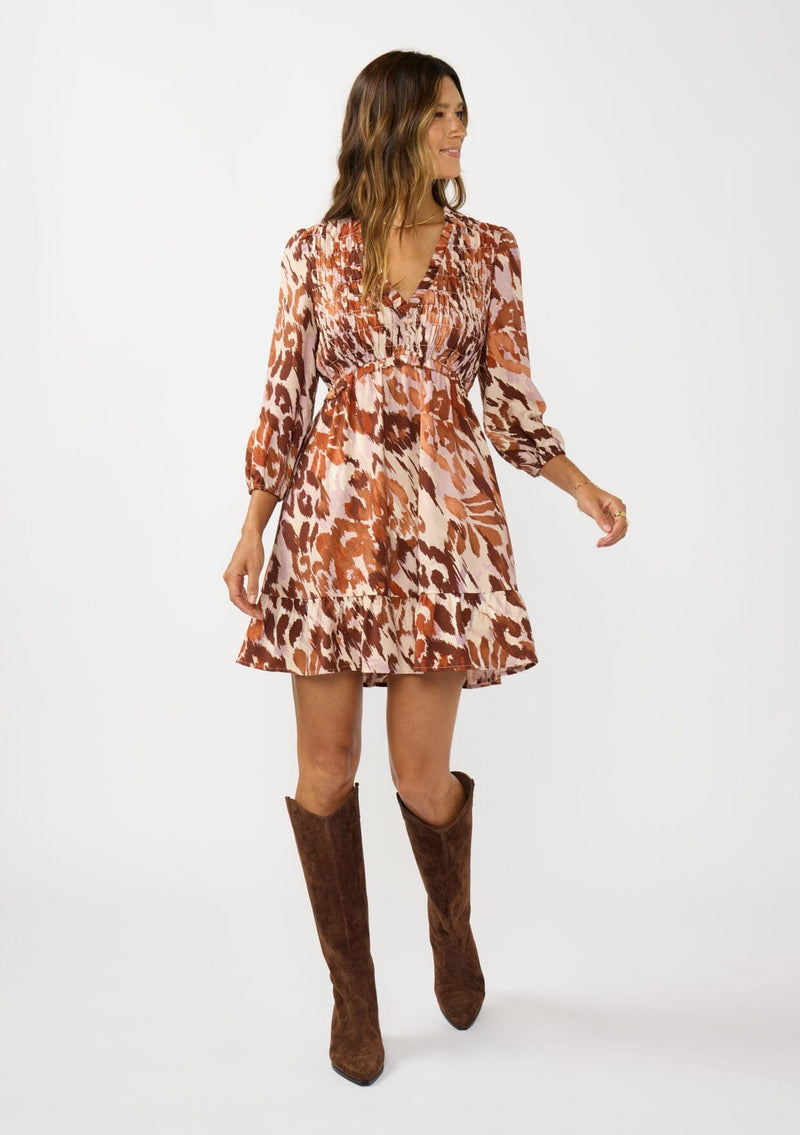 [Color: Nude/Brown] A front facing image of a brunette model wearing a brown abstract print mini dress with a smocked top, long sleeves, and a ruffle trimmed hem. A fall boho dress for casual outings. 