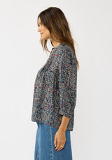 [Color: Navy/Rose] A side facing image of a brunette model wearing a navy floral blouse with 3/4 length sleeves, a split v-neckline, and front pintuck details. A classic boho blouse for casual outings. 
