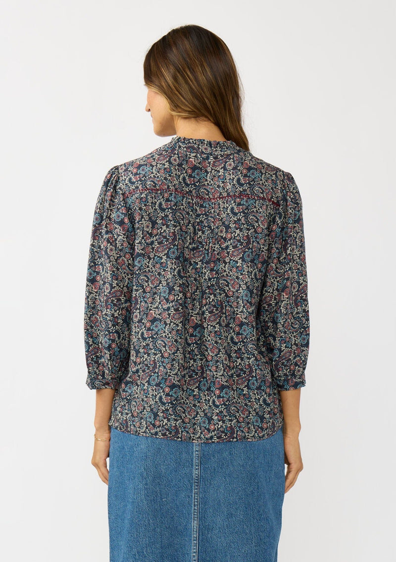 [Color: Navy/Rose] A back facing image of a brunette model wearing a navy floral blouse with 3/4 length sleeves, a split v-neckline, and front pintuck details. A classic boho blouse for casual outings. 