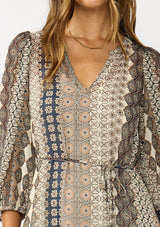 [Color: Natural/Navy] A detail facing image of a brunette model wearing boho floral maxi dress with 3/4 volume sleeves, waist belt, and a high low tiered skirt. 