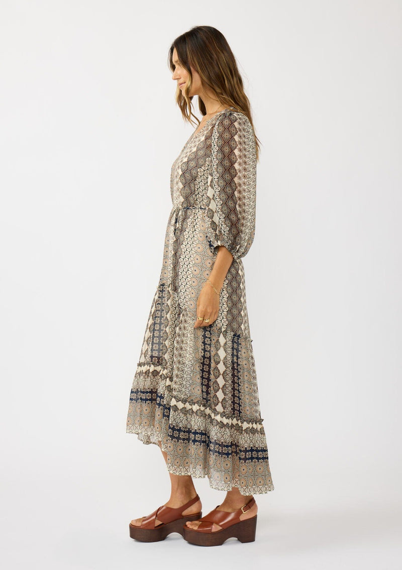 [Color: Natural/Navy] A side facing image of a brunette model wearing boho floral maxi dress with 3/4 volume sleeves, waist belt, and a high low tiered skirt. 