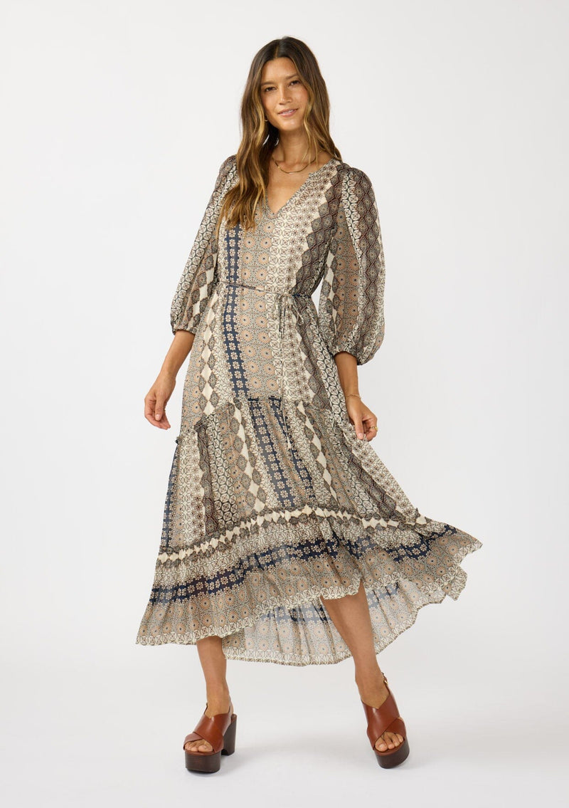 [Color: Natural/Navy] A front facing image of a brunette model wearing boho floral maxi dress with 3/4 volume sleeves, waist belt, and a high low tiered skirt. 