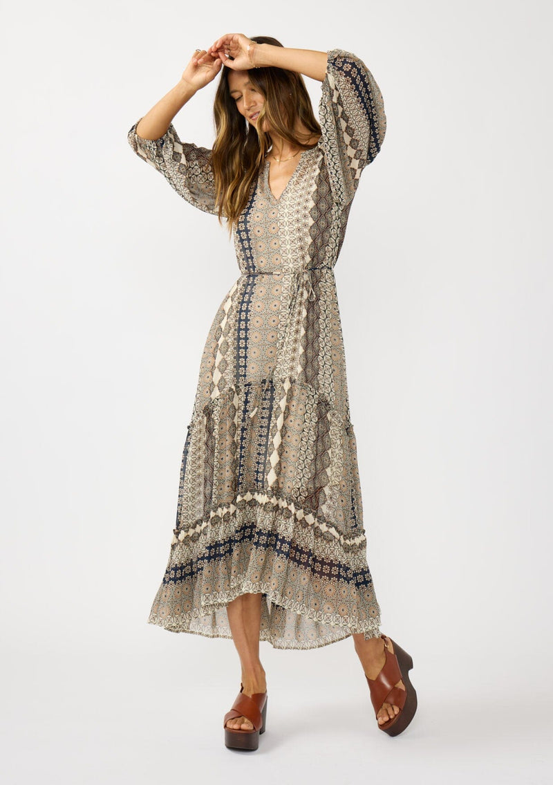 [Color: Natural/Navy] A front facing image of a brunette model wearing boho floral maxi dress with 3/4 volume sleeves, waist belt, and a high low tiered skirt. 