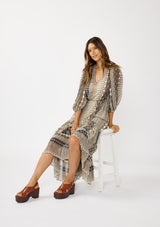 [Color: Natural/Navy] A sitting image of a brunette model wearing boho floral maxi dress with 3/4 volume sleeves, waist belt, and a high low tiered skirt. 