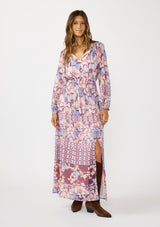 [Color: Natural/Dusty Blue] A front facing image of a brunette model wearing a blue and pink floral maxi dress with long sleeves, a split v-neckline with ties, a smocked waist, and a side slit. 