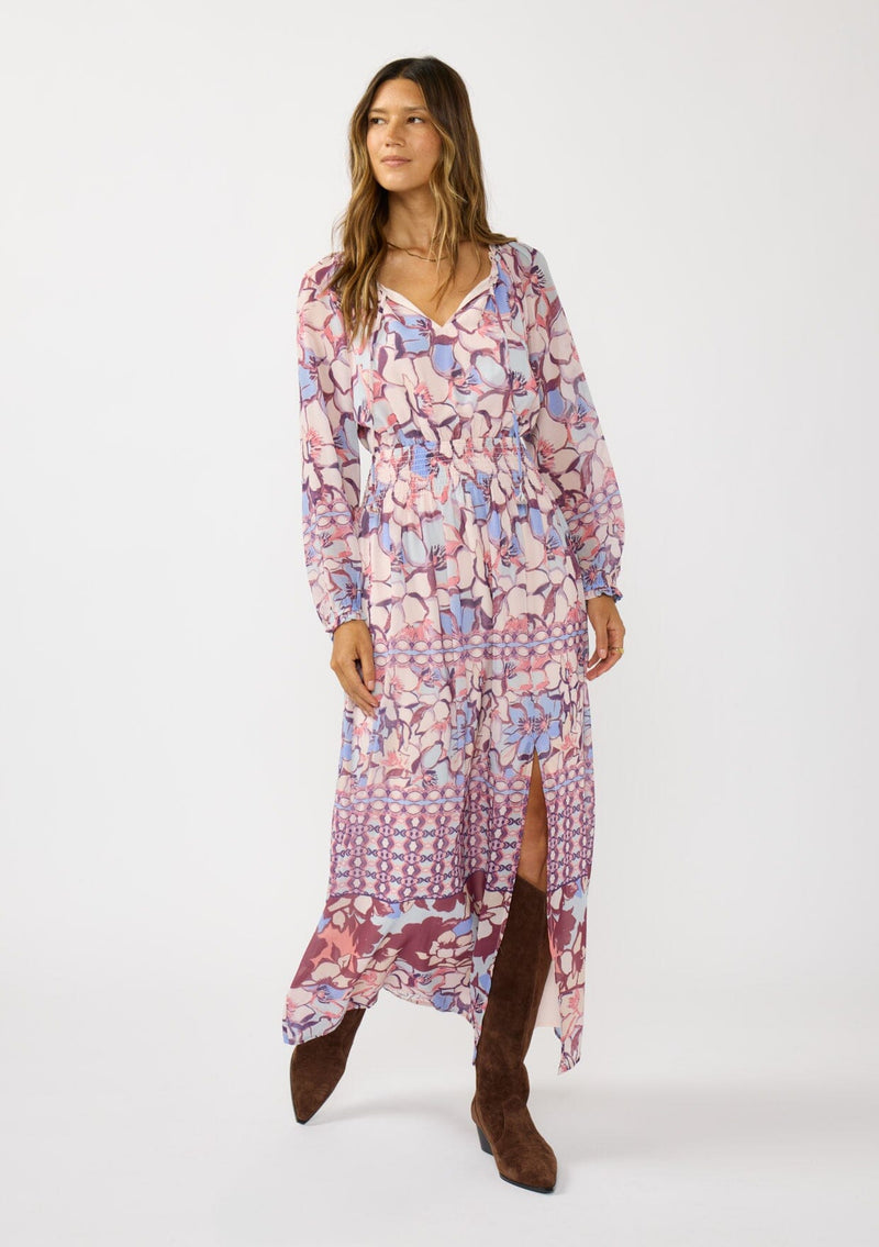 [Color: Natural/Dusty Blue] A front facing image of a brunette model wearing a blue and pink floral maxi dress with long sleeves, a split v-neckline with ties, a smocked waist, and a side slit. 