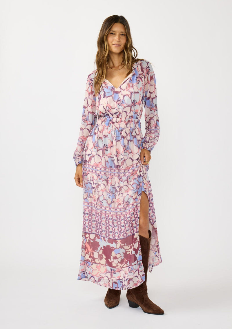 [Color: Natural/Dusty Blue] A front facing image of a brunette model wearing a blue and pink floral maxi dress with long sleeves, a split v-neckline with ties, a smocked waist, and a side slit. 