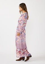 [Color: Natural/Dusty Blue] A side facing image of a brunette model wearing a blue and pink floral maxi dress with long sleeves, a split v-neckline with ties, a smocked waist, and a side slit. 